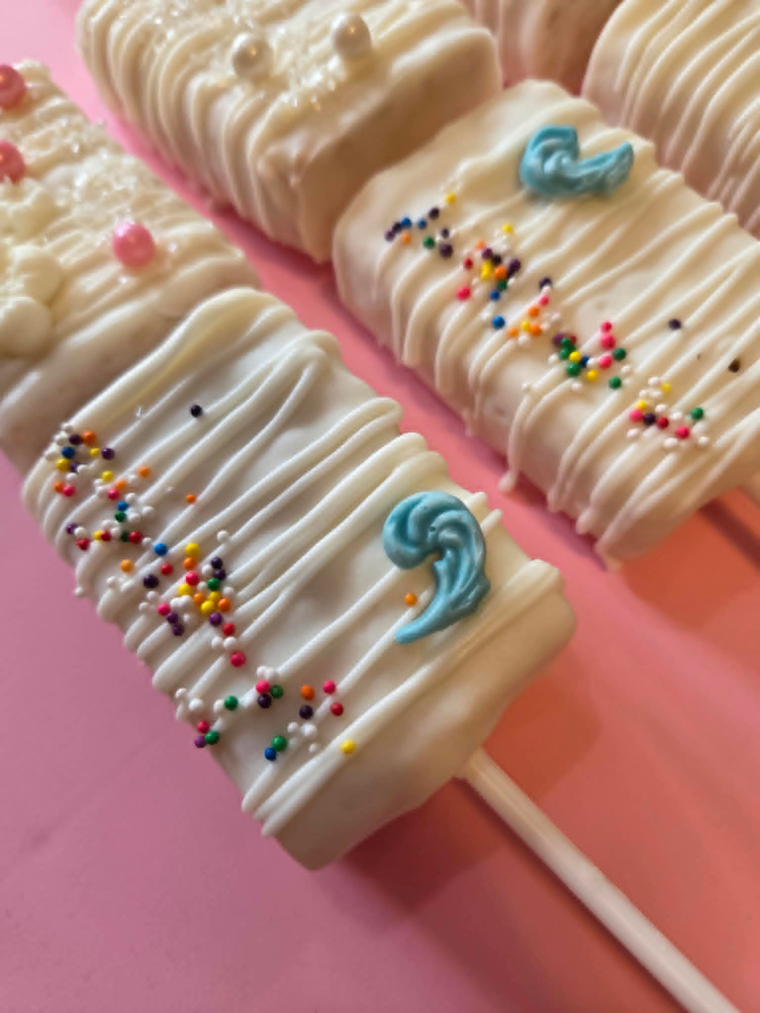 White chocolate Oreo pops with rainbow sprinkles and blue wave toppers on pink background