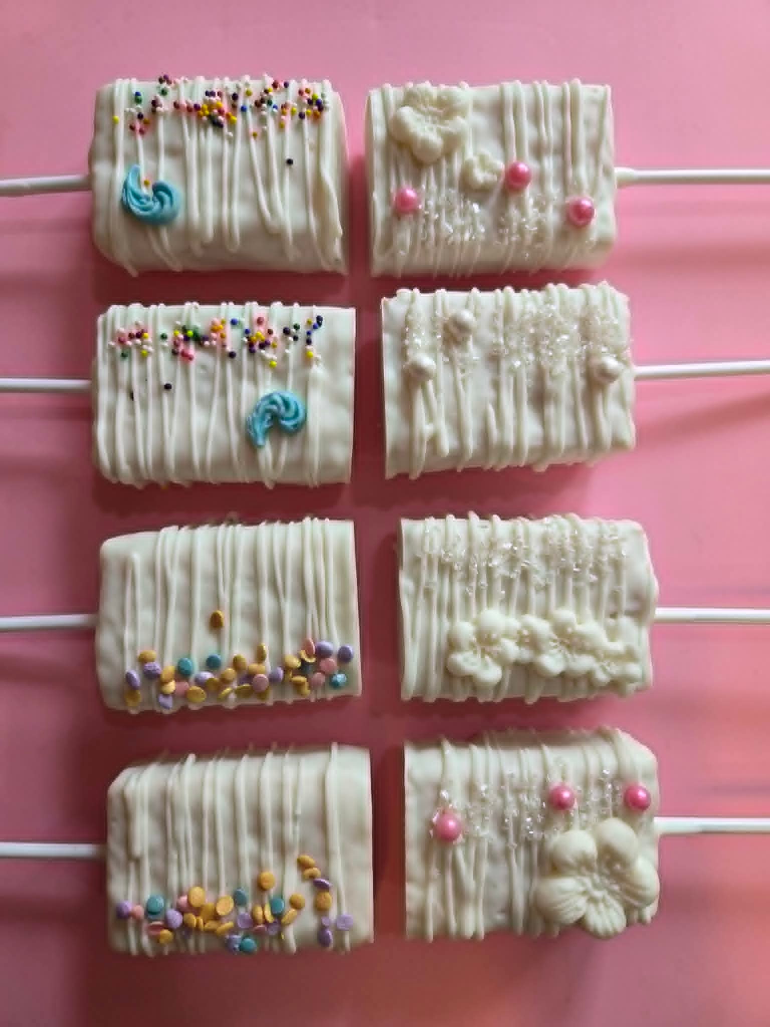 White chocolate rice krispy treats on sticks with colorful sprinkles and flower toppers on pink background
