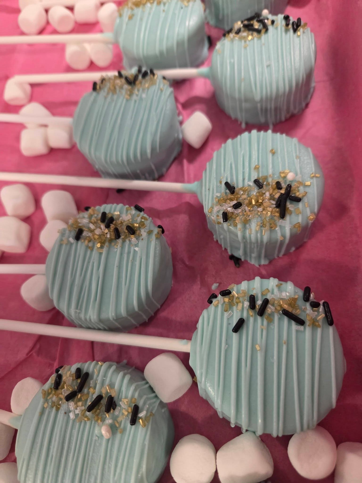 Teal chocolate-dipped Oreo pops with gold and black sprinkles on pink background